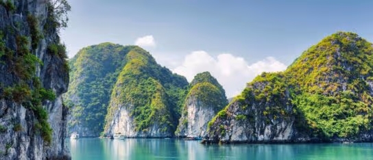 Lush green limestone karst islands rise steeply out of calm emerald bay water under a partly cloudy sky.