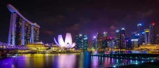Illuminated skyscrapers and modern architecture along a bay in Singapore at night with colourful reflections on the water.