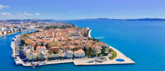 Aerial view of a coastal city peninsula with red-roofed buildings surrounded by blue ocean water under a clear sky.