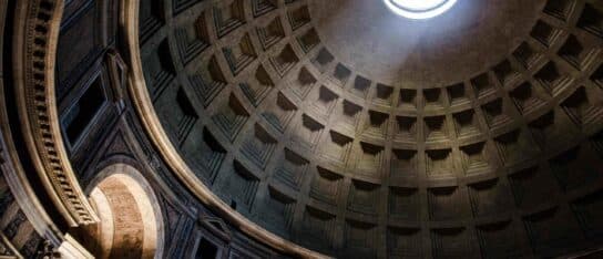 Low-angle view looking up at a coffered stone dome with light streaming through a central circular opening.