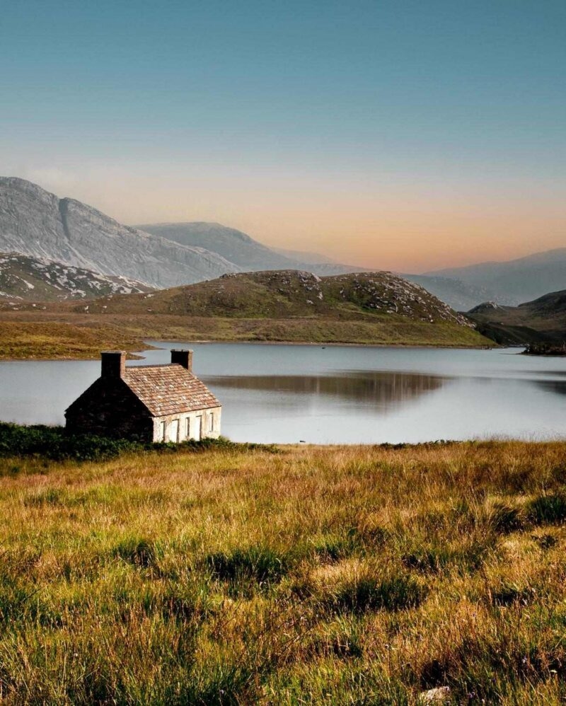 Small stone cottage beside a quiet lake in a grassy valley with mountain peaks in the distance.