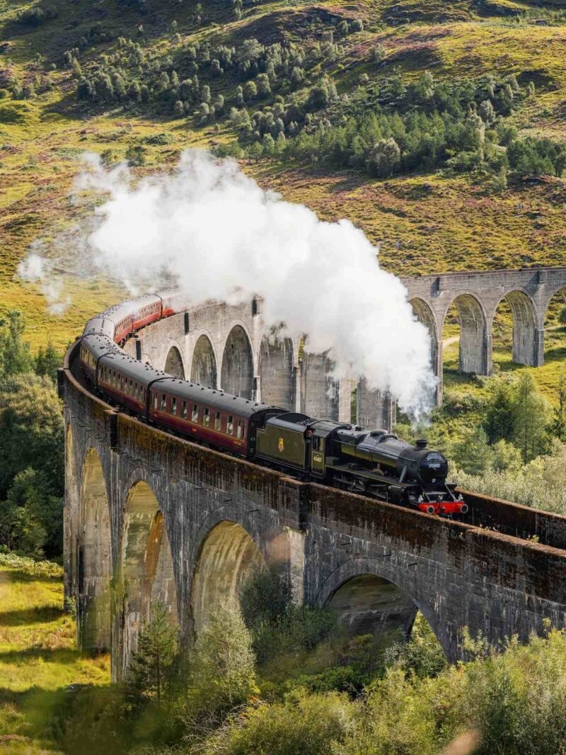 A vintage steam train emitting white smoke crosses a stone arch bridge in a mountainous landscape.