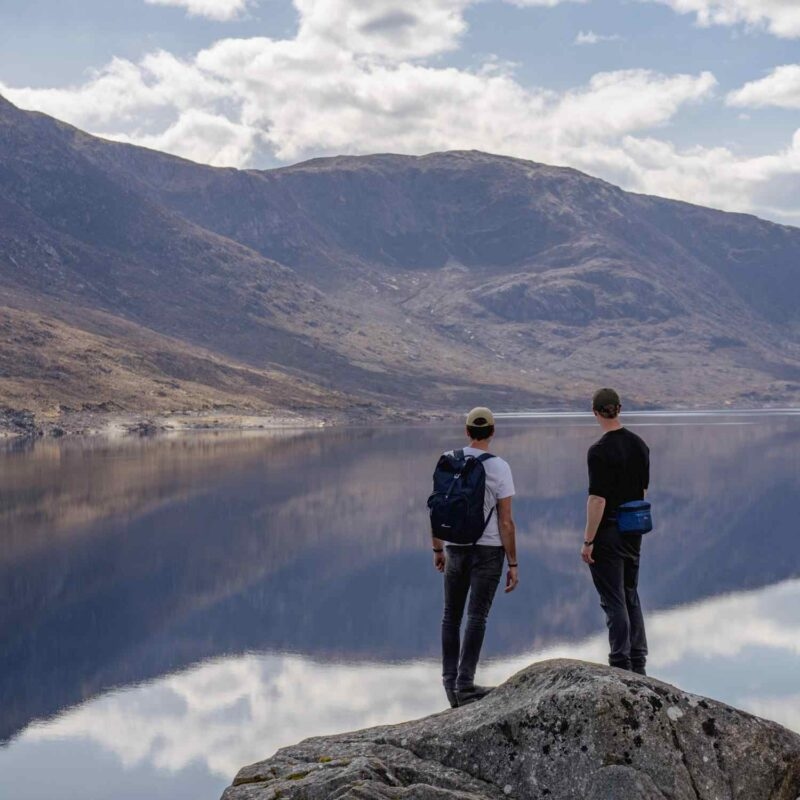 Two hikers on a rock look out at a tranquil lake reflecting mountains and clouds in the background.