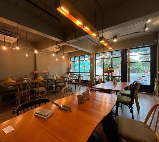 Cosy interior of a cafe with wooden tables, chairs, hanging warm lights, and glass windows leading outside.