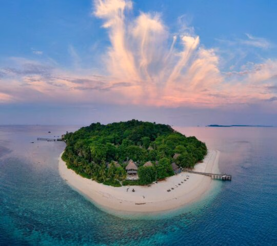 An aerial view of a small island surrounded by the ocean, with pale sandy beaches and a densely forested interior, and a jetty visible to the right. You can see the roofs of villas through the trees.