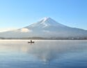 A small boat on a misty lake reflecting a large snow-capped mountain under a clear blue sky.