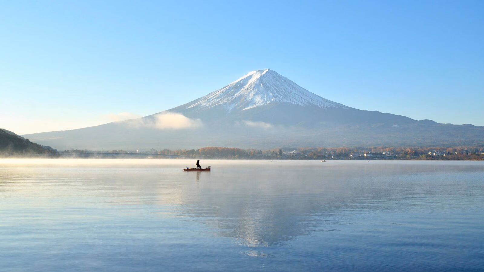 A small boat on a misty lake reflecting a large snow-capped mountain under a clear blue sky.