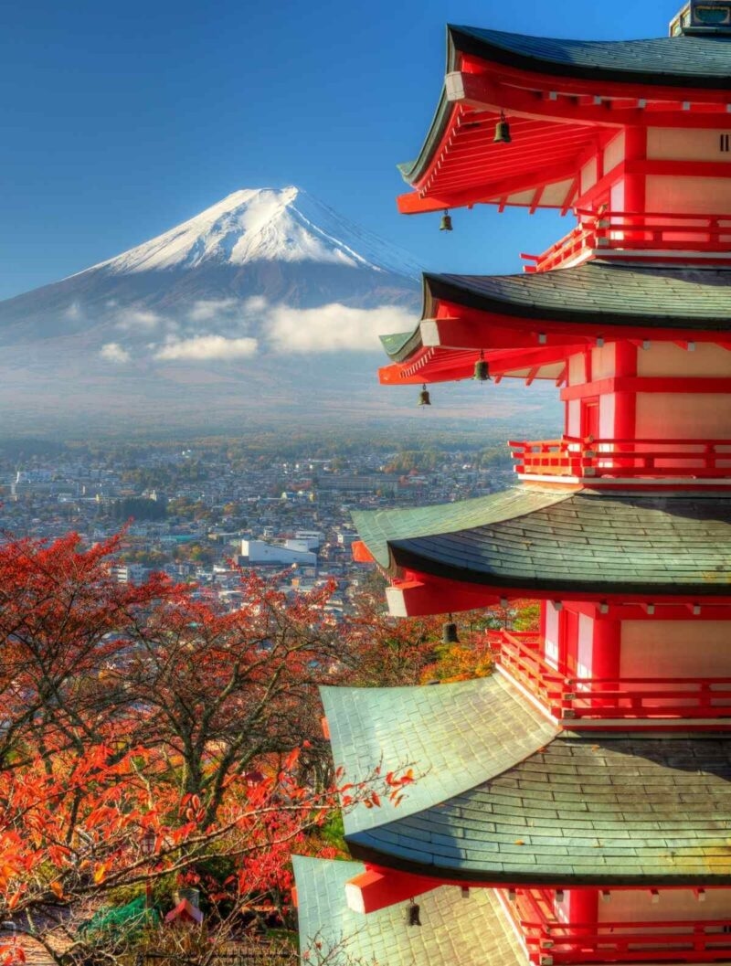 Traditional red pagoda overlooking a city with autumn trees and a snow-capped mountain in the distance.