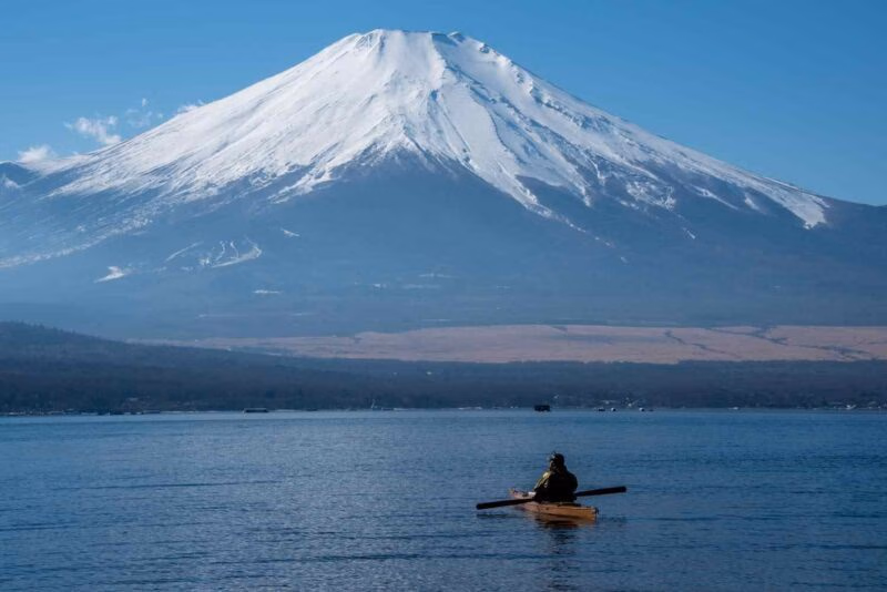 Kayaker on a calm lake in front of a massive snow-capped mountain under a clear blue sky.