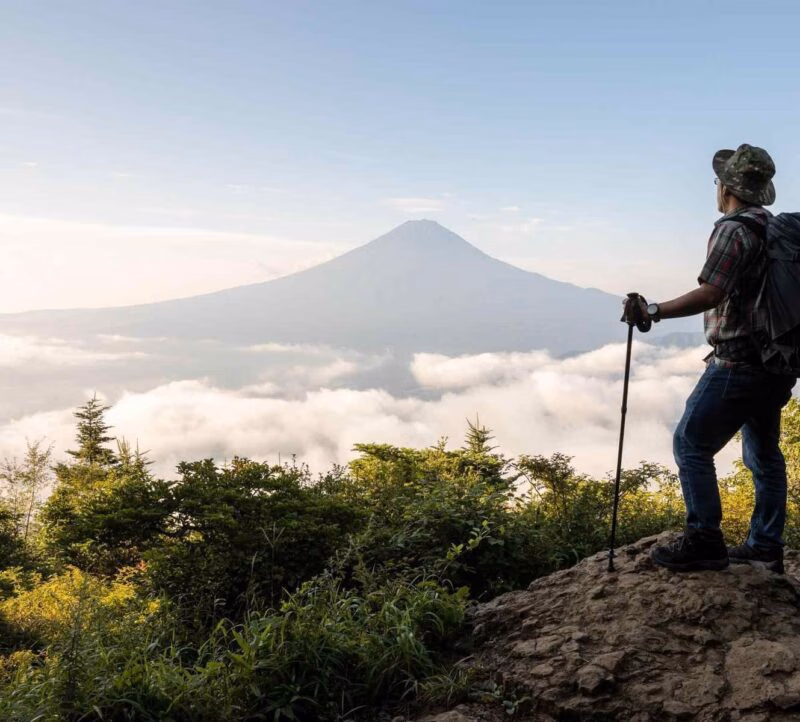 Hiker with a backpack and trekking pole standing on a rock looking at a distant mountain peak above clouds.