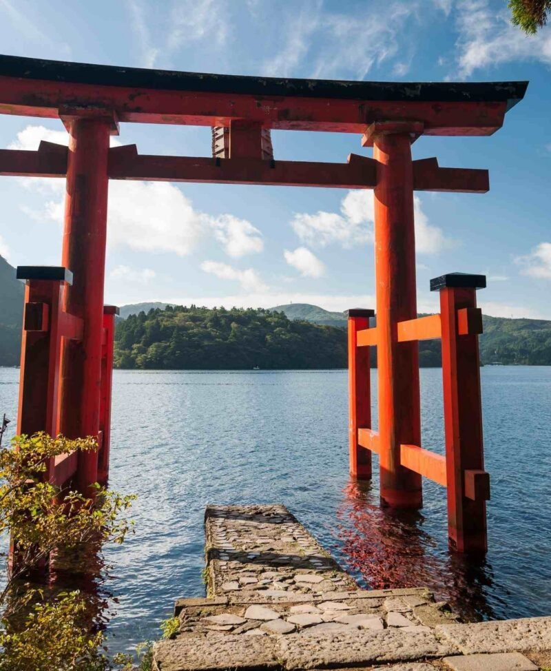 Large red torii gate in blue water with a stone pathway leading toward forest-covered hills.