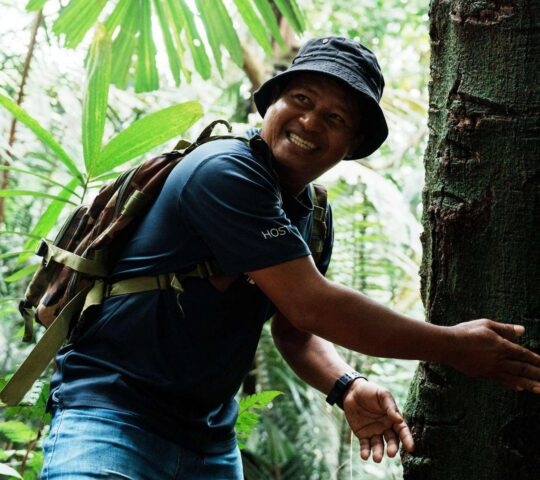 A man wearing a black bucket hat and backpack smiles as he touches a tree trunk in a dense forest.
