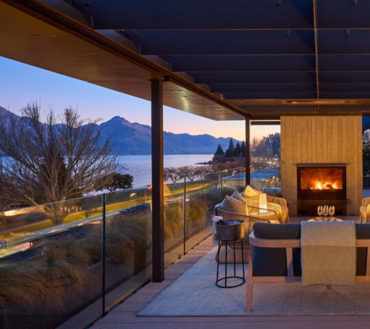 Side view of a lounge area with sofas and armchairs in front of a wood-burning stove, under a wooden roof with beams and looking out over a glass balcony at lake and mountain views.