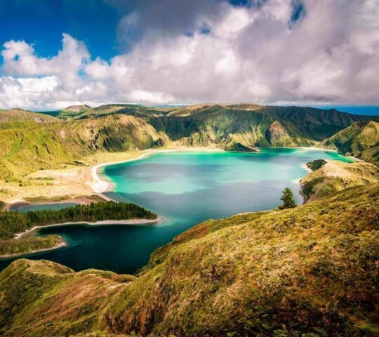 Elevated landscape view of a turquoise crater lake surrounded by green sloped hills under a partially cloudy sky.
