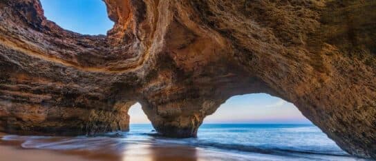 Interior view of a sea cave with natural rock arches opening onto a sandy beach and blue ocean.
