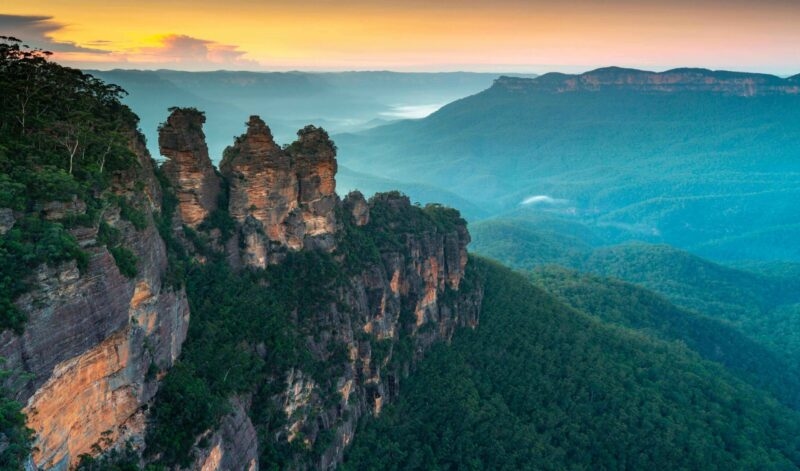 The Three Sisters rock formation rising over dense green forest and misty mountain valleys at sunrise.