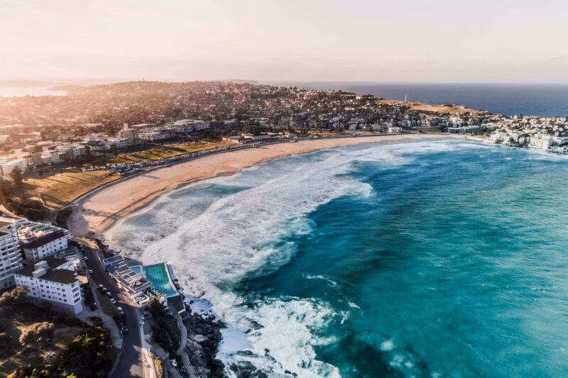 Aerial shot of Bondi Beach in Australia with turquoise ocean waves rolling onto a sandy shore next to coastal buildings.