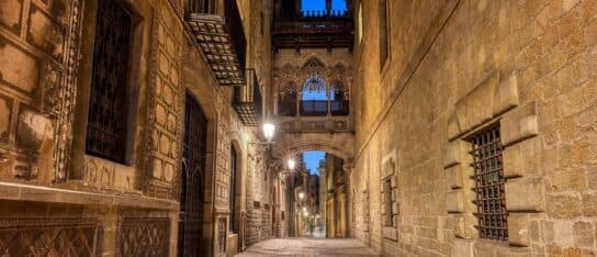 An ornate gothic bridge connects stone buildings over an illuminated cobblestone alleyway at dusk.