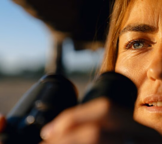 Close-up of a woman's face as she holds up binoculars in warm sunlight.