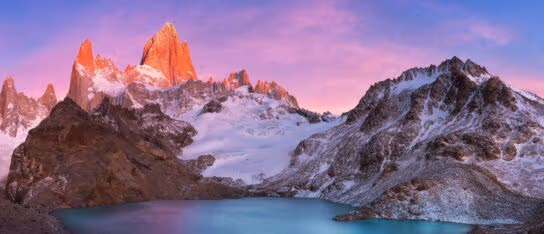 Snow-capped jagged mountain peaks illuminated by pink sunrise light over a calm blue lake.