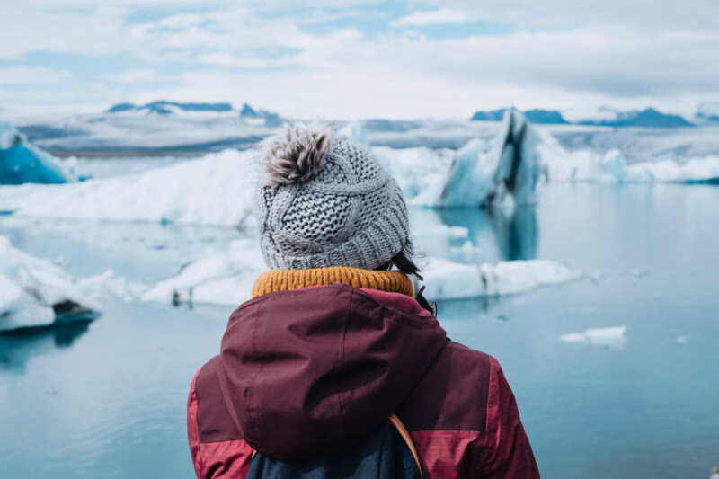 Person in a knit hat looking at large blue icebergs floating in a glacial lagoon on a polar safari.