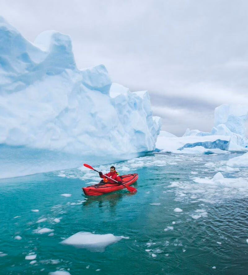 Person kayaking in a red kayak on bright turquoise water near massive icebergs during a polar safari.