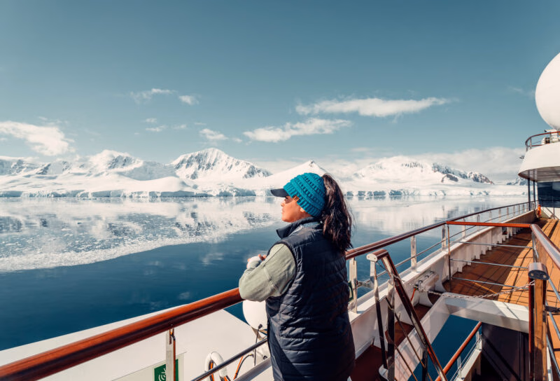 Woman in a teal hat standing on a ship deck looking at snow-covered mountains on a polar safari.