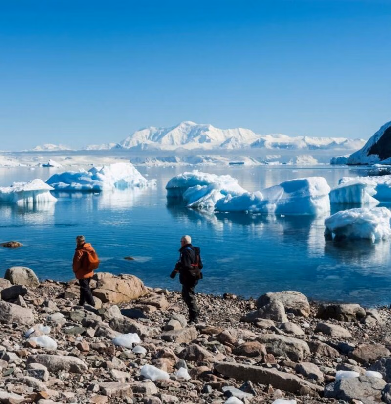 Two people walking on a rocky shore looking at floating icebergs and distant snowy peaks on a polar safari.