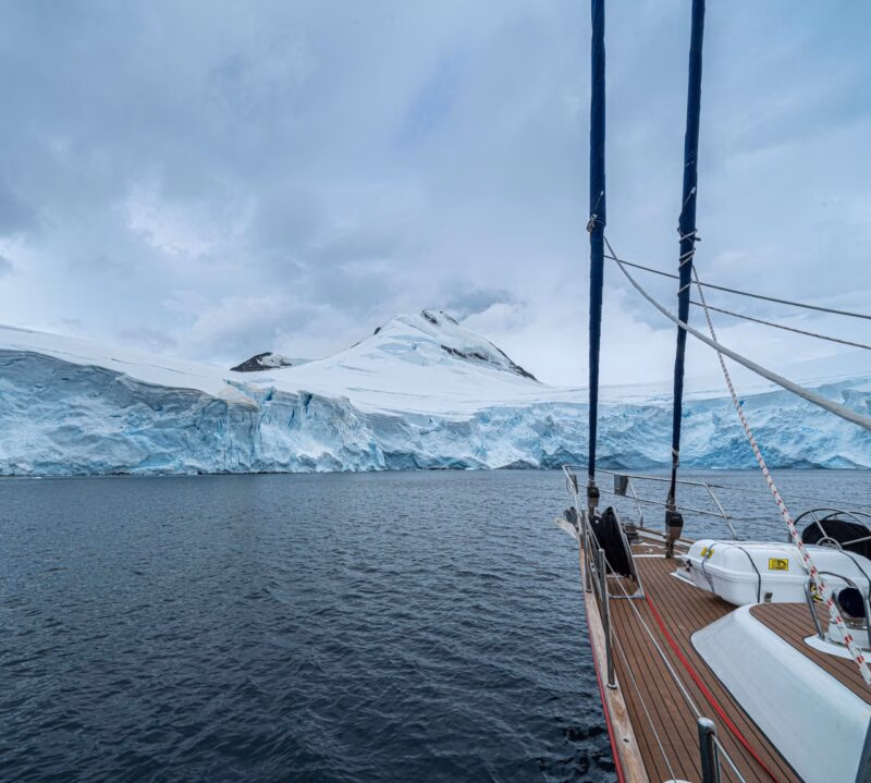 The bow of a sailing yacht approaches a colossal, towering glacier wall rising from the dark water during an exploratory polar safari.