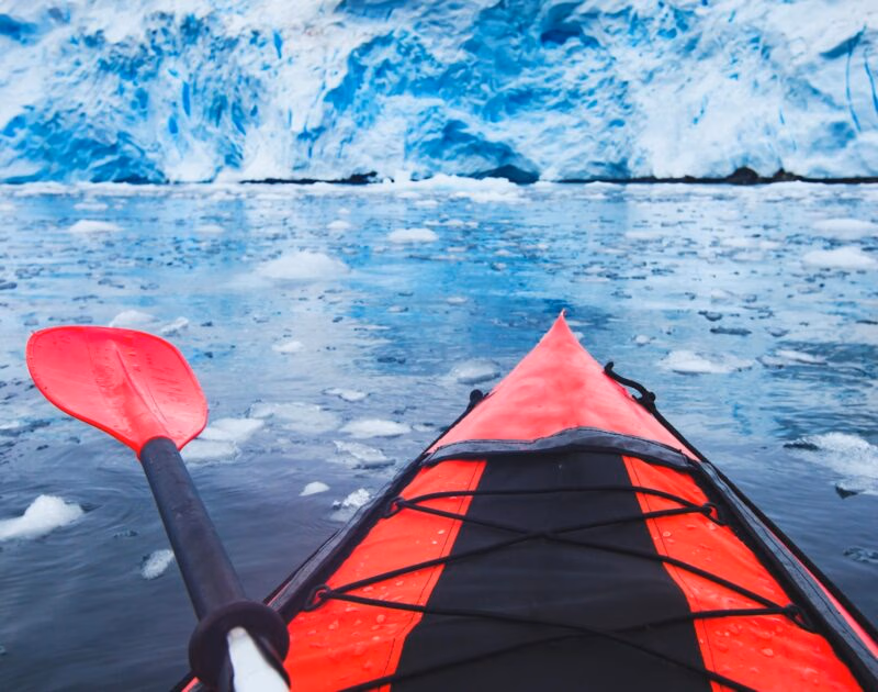 First-person view from a red kayak facing a massive blue ice wall in icy water on a polar safari.