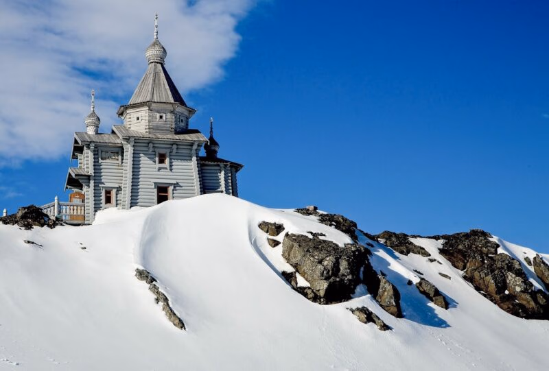 Wooden church with traditional onion domes perched on a snowy, rocky hill under a blue sky on a polar safari.