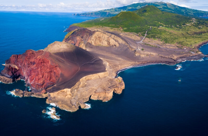 Aerial view of a red and brown volcanic peninsula meeting the blue ocean, with green mountains inland.