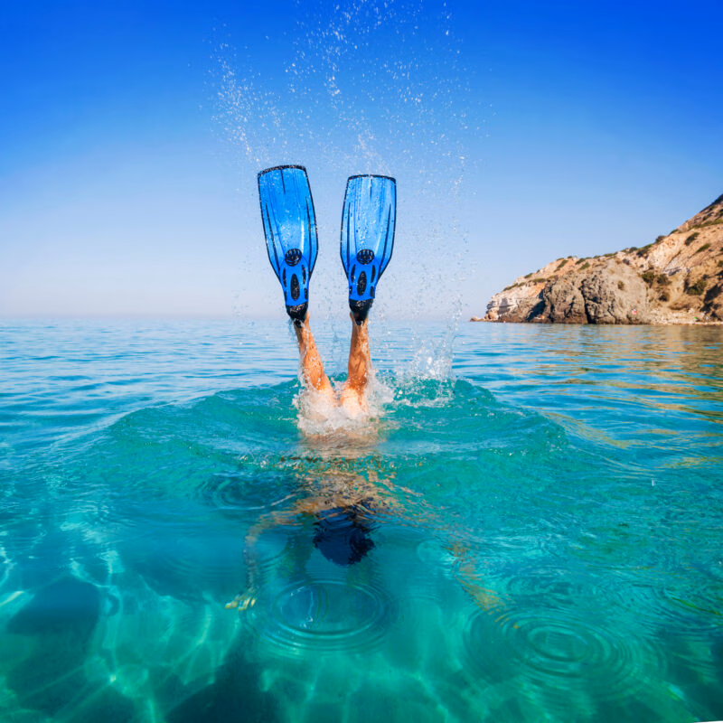 Blue snorkeling fins splashing above bright turquoise water as a person dives under, with a rocky coast behind.
