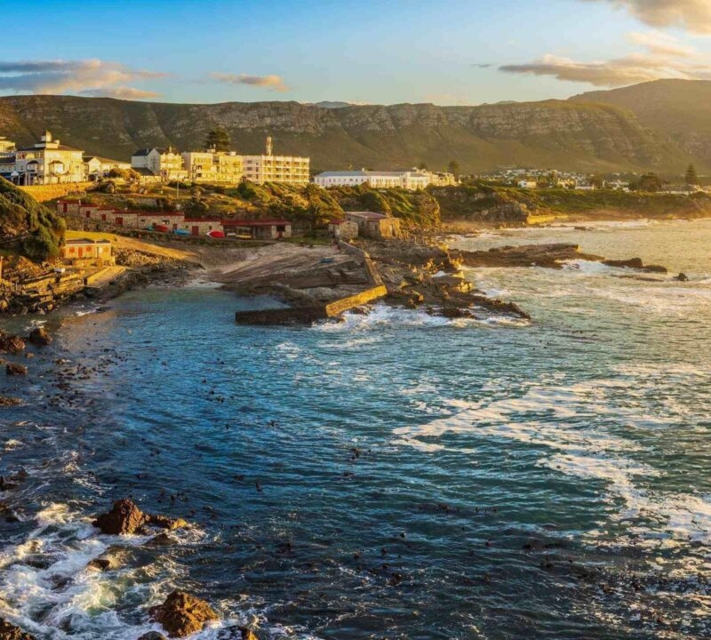 Coastal town buildings on a cliff at sunset with crashing ocean waves on rocky shores and distant mountains.