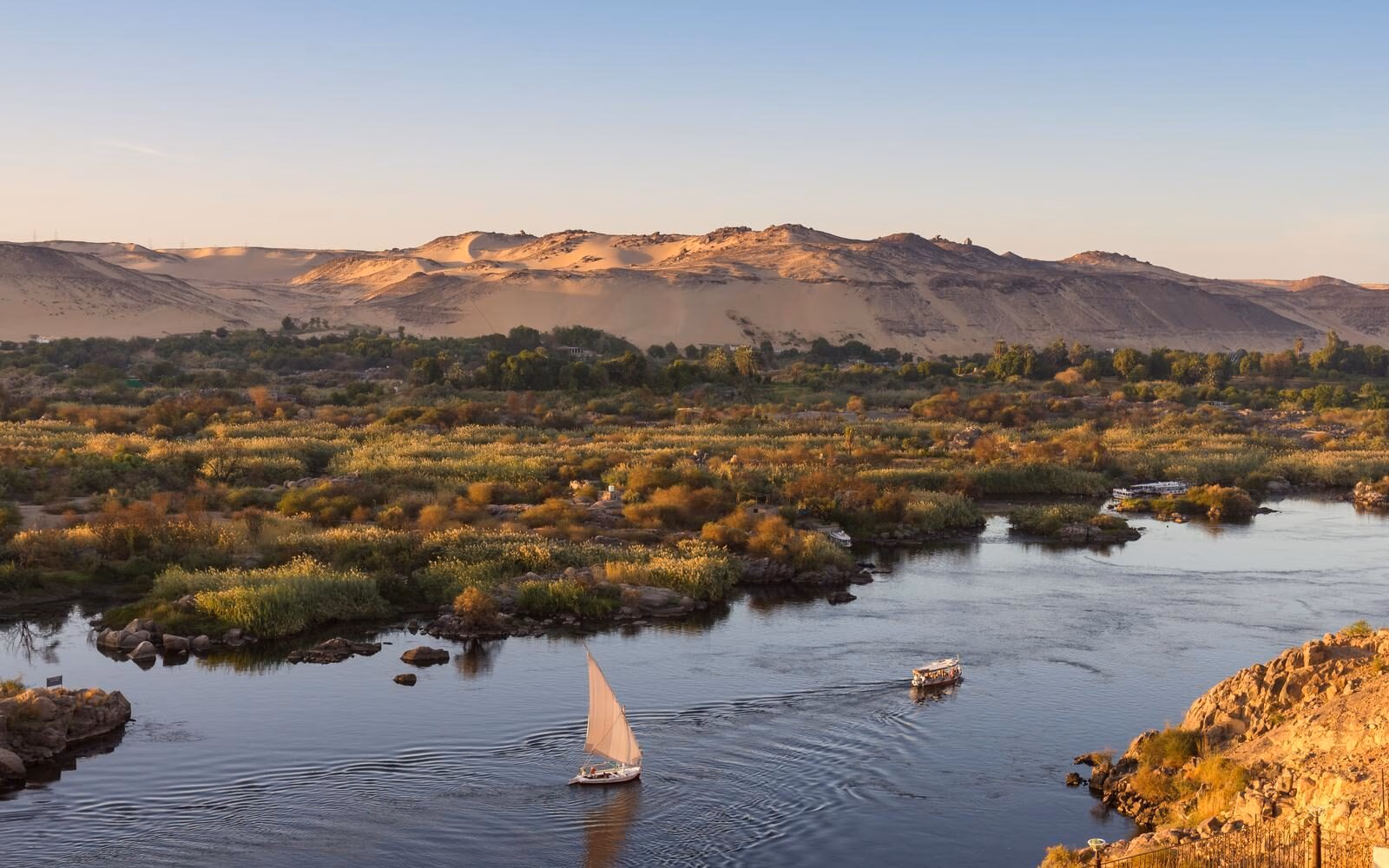 Sail along the Nile in a traditional felucca, passing lush riverbanks and golden desert hills around Aswan, Egypt.