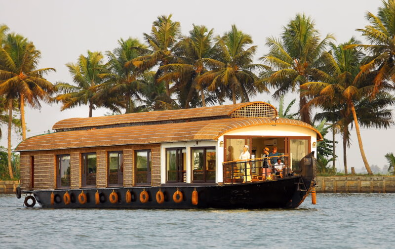 A houseboat on a Spice Routes Cruise sailing along the Keralan Backwaters in India, surrounded by palm trees.