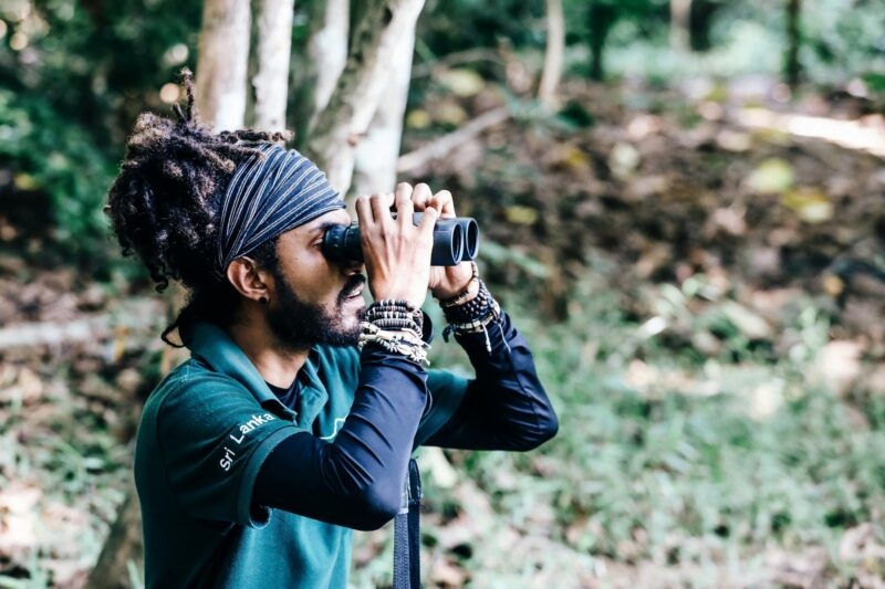 A man wearing a green shirt with the text Sri Lanka looks through binoculars in a forest setting.