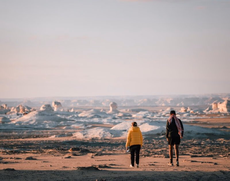 Two people walking away across a sandy Egypt desert landscape with large white rock formations under a pale sky.