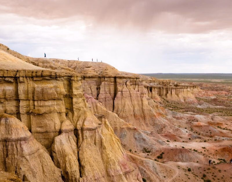 People stand on colorful eroded cliffs in the gobi desert mongolia under a cloudy sky.