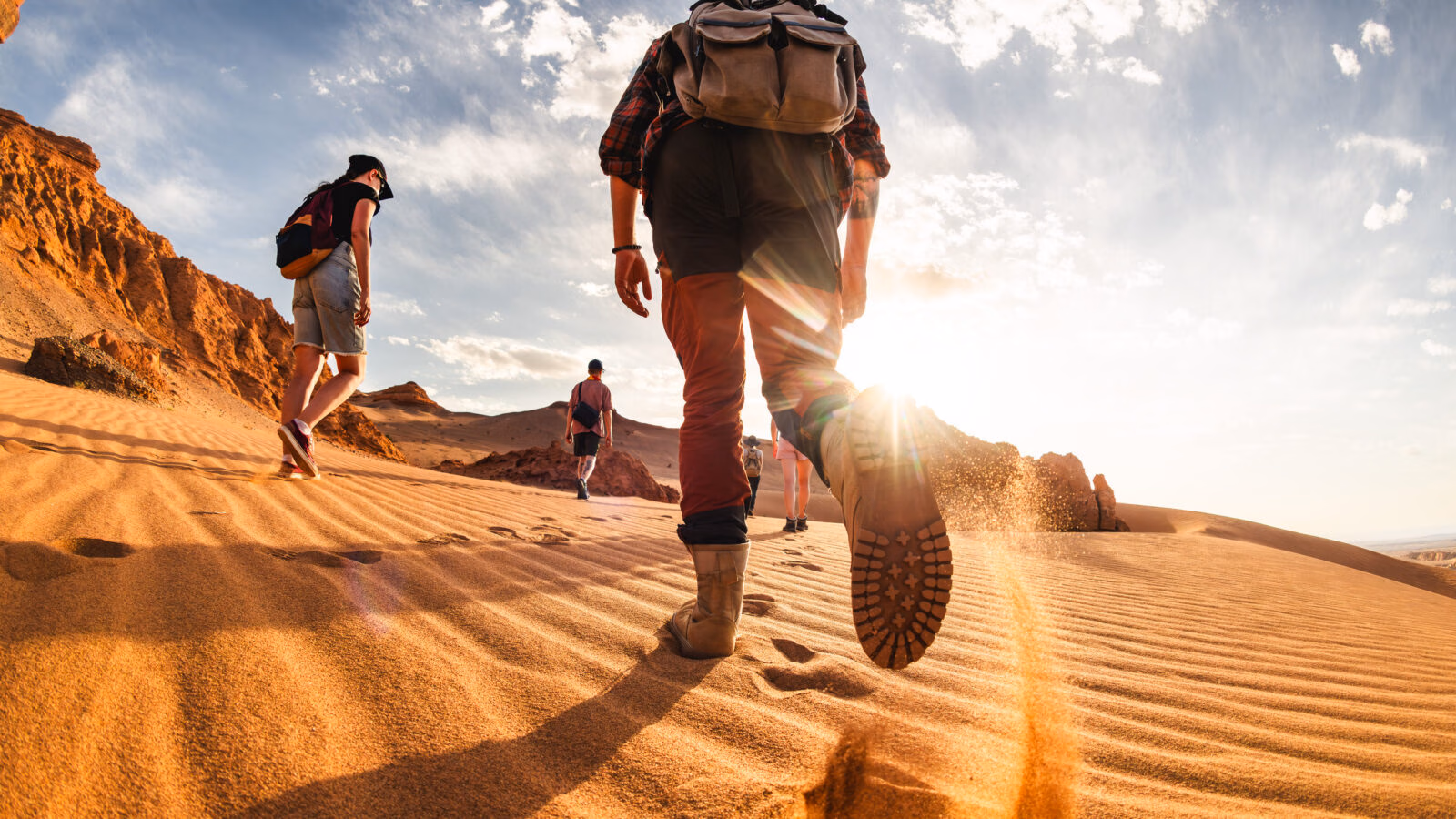 A group of hikers walking across golden sand dunes in Mongolia’s Gobi Desert, surrounded by rocky formations in the afternoon sunlight.