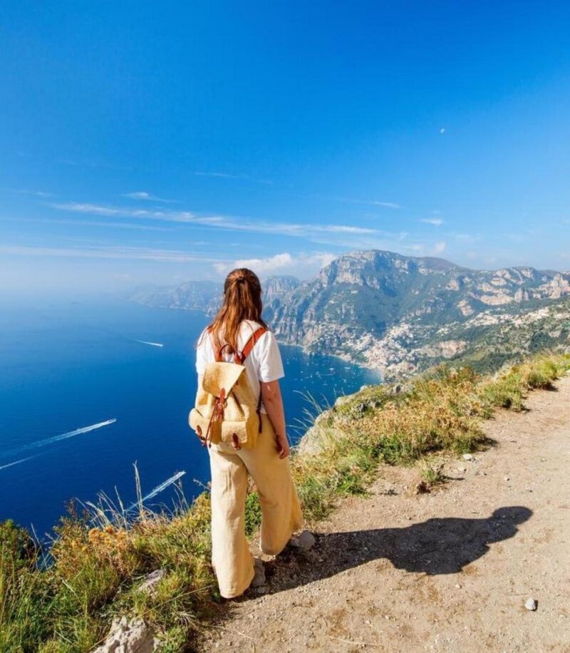 A woman with a backpack on a high cliff trail overlooking the blue sea and a coastal town.