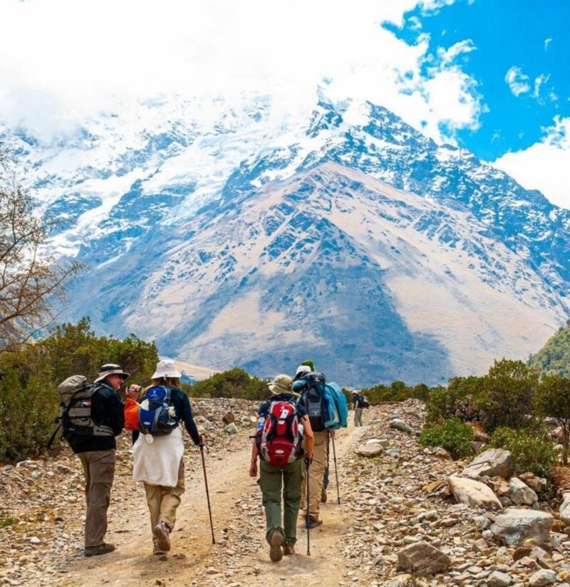 Hikers with backpacks walk a rocky trail toward a massive snow-capped mountain under a bright blue sky.