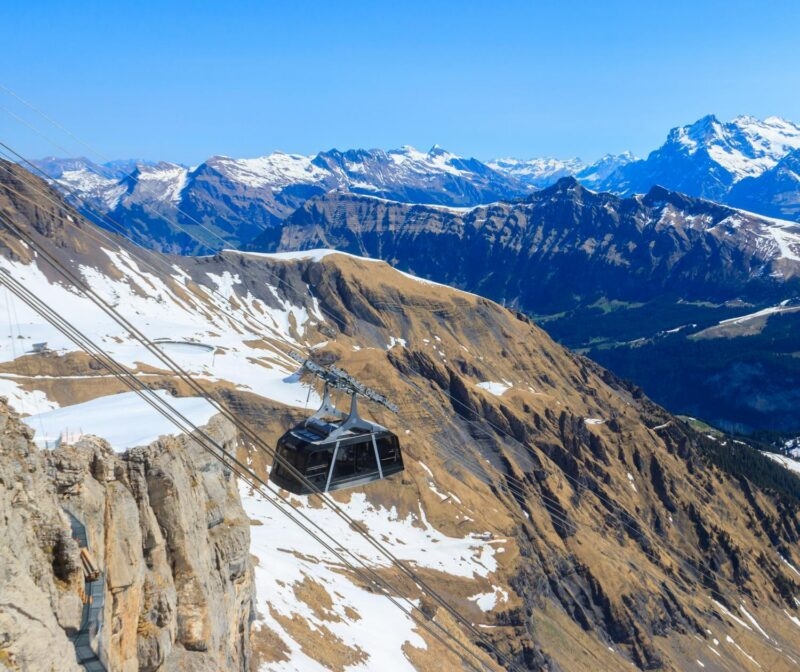 A black cable car swiss travels on wires over steep, partially snow-covered mountains under a clear blue sky