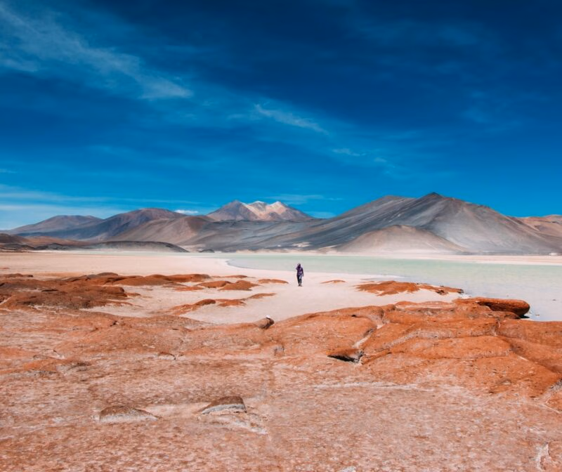 Person walking near red rock formations and a turquoise lagoon with mountains under a deep blue sky.