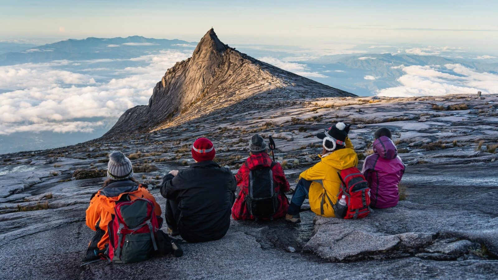 Five hikers sit on a rocky slope looking at a sharp mountain peak surrounded by clouds.