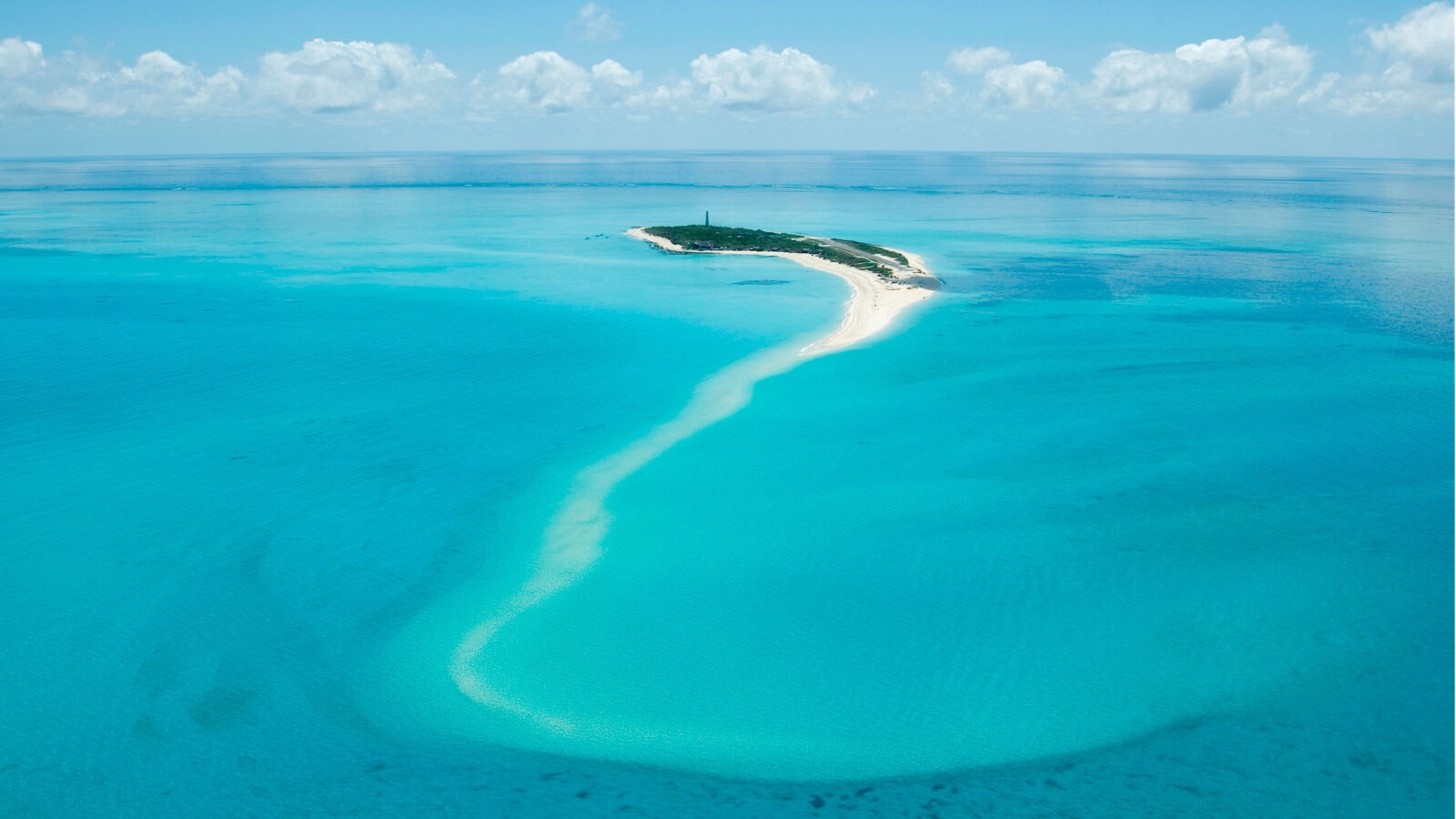 Aerial view of a small green island with a long, winding white sandbar extending into clear turquoise water.