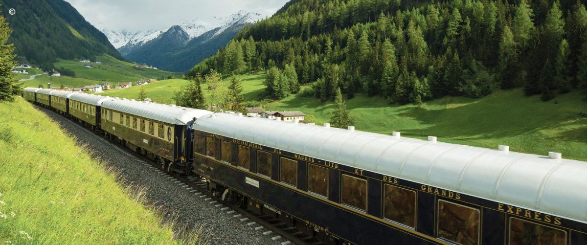 The Venice Simplon Orient Express travelling through the lush green countryside bordered by snowcapped mountains