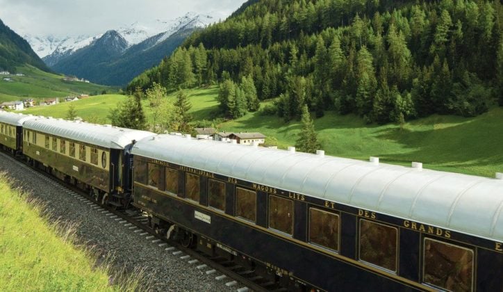 The Venice Simplon Orient Express travelling through the lush green countryside bordered by snowcapped mountains