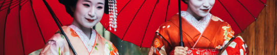 Two maiko geisha walking on a street in Kyoto with red umbrellas