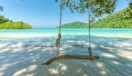 A swing on a beach in Thailand with an idyllic view of the water and lush hills.